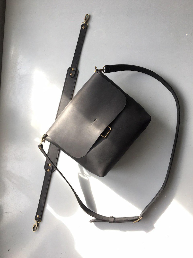 Puede incluir: Bolso bandolera de cuero gris oscuro con cierre de solapa y correa de hombro larga y ajustable. El bolso presenta herrajes dorados y una correa desmontable. El dise&ntilde;o es minimalista y moderno.