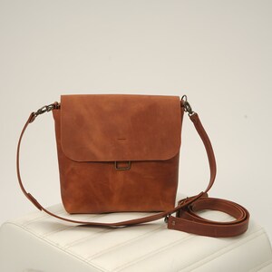 Puede incluir: Bolso bandolera de cuero marr&oacute;n con cierre de solapa y correa ajustable para el hombro. El bolso tiene forma rectangular y un cierre met&aacute;lico. La correa est&aacute; sujeta con herrajes met&aacute;licos. El bolso est&aacute; sobre una superficie blanca.