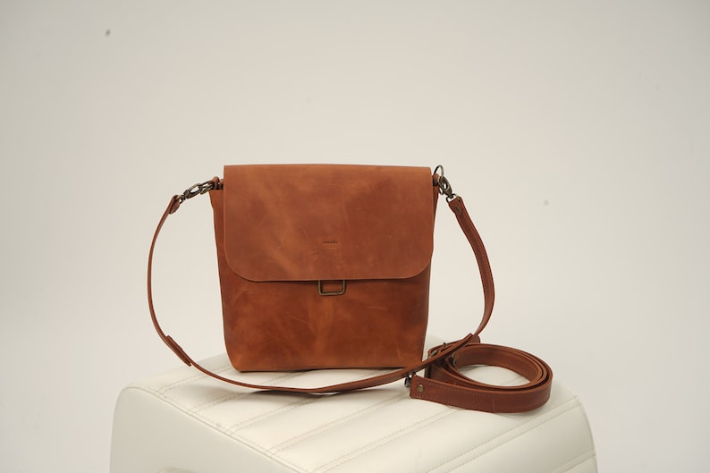 Puede incluir: Bolso bandolera de cuero marr&oacute;n con cierre de solapa y correa ajustable para el hombro. El bolso tiene forma rectangular y un cierre met&aacute;lico. La correa est&aacute; sujeta con herrajes met&aacute;licos. El bolso est&aacute; sobre una superficie blanca.