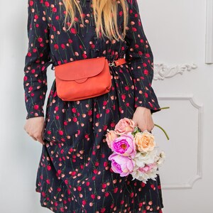 May include: A woman wearing a navy blue dress with a red floral pattern, a matching orange belt bag, and holding a bouquet of pink, peach, and white flowers. The dress has long sleeves.
