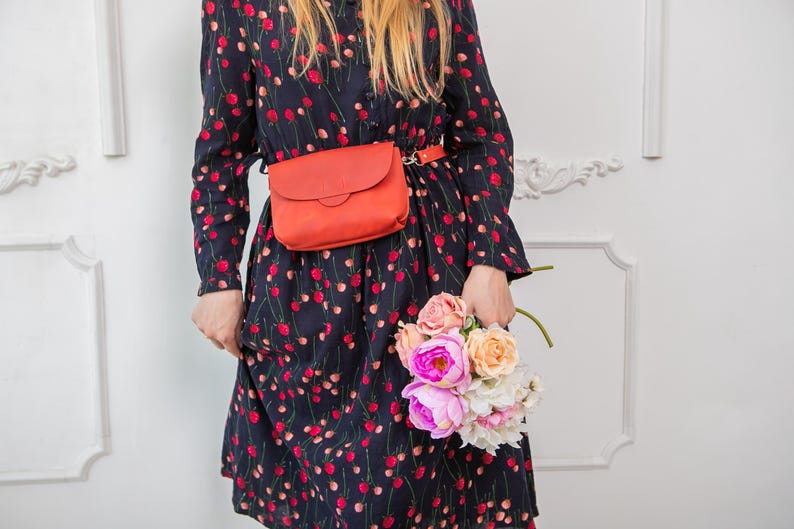 May include: A woman wearing a navy blue dress with a red floral pattern, a matching orange belt bag, and holding a bouquet of pink, peach, and white flowers. The dress has long sleeves.