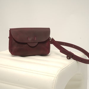 May include: A burgundy leather crossbody bag with a flap closure and a matching adjustable strap. The bag features a rounded flap with decorative stitching and a rectangular body. The bag is displayed on a white surface.