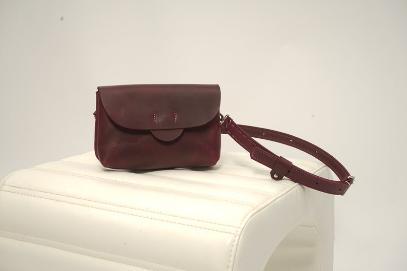 May include: A burgundy leather crossbody bag with a flap closure and a matching adjustable strap. The bag features a rounded flap with decorative stitching and a rectangular body. The bag is displayed on a white surface.