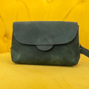 May include: A dark green leather crossbody bag with a flap closure and a matching adjustable strap. The bag has a rounded flap and a small, circular clasp. The bag is set against a bright yellow background.
