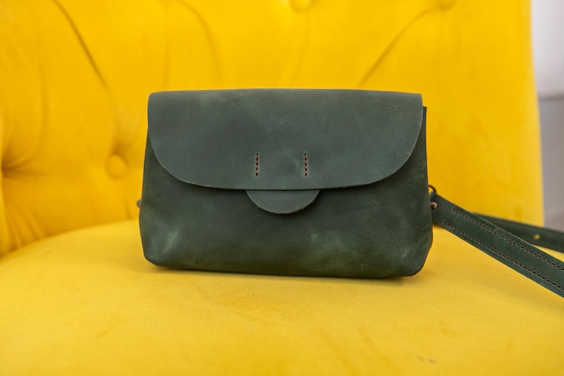 May include: A dark green leather crossbody bag with a flap closure and a matching adjustable strap. The bag has a rounded flap and a small, circular clasp. The bag is set against a bright yellow background.