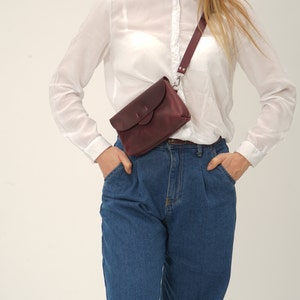 May include: A burgundy leather crossbody bag with a flap closure and adjustable strap. The bag is worn across the body, paired with blue jeans and a white button-down shirt. The bag has a silver-tone clasp.