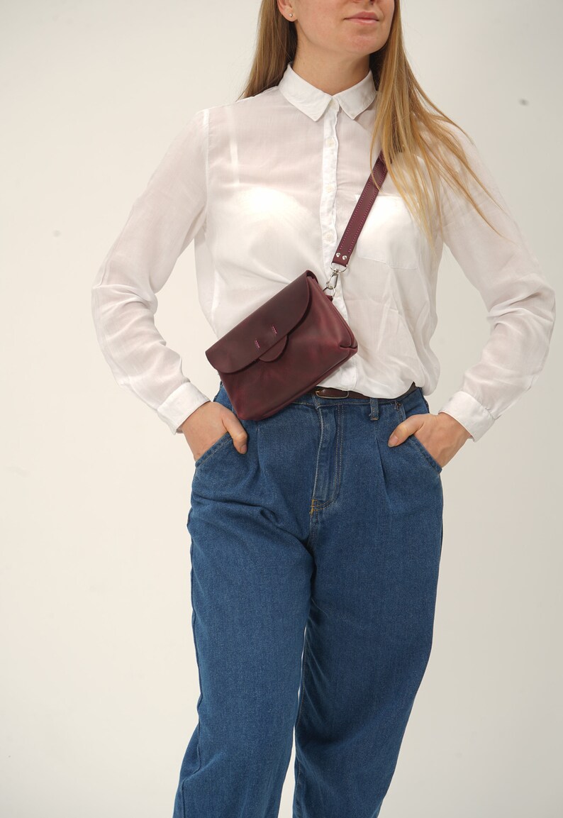 May include: A burgundy leather crossbody bag with a flap closure and adjustable strap. The bag is worn across the body, paired with blue jeans and a white button-down shirt. The bag has a silver-tone clasp.