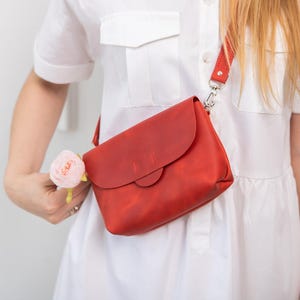 May include: A red leather crossbody bag with a flap closure and adjustable strap. The bag is worn over the shoulder. The bag is a vibrant red color and appears to be made of high-quality leather.