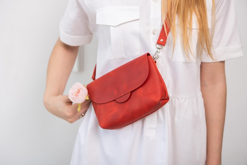 May include: A red leather crossbody bag with a flap closure and adjustable strap. The bag is worn over the shoulder. The bag is a vibrant red color and appears to be made of high-quality leather.