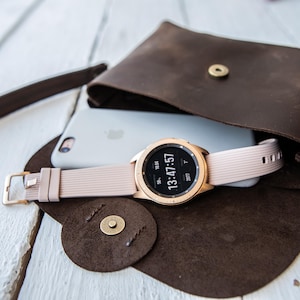 May include: A rose gold smartwatch with a light pink band rests on a white phone inside a brown leather crossbody bag. The watch displays the time 13:47:57. The bag is open, revealing its contents.