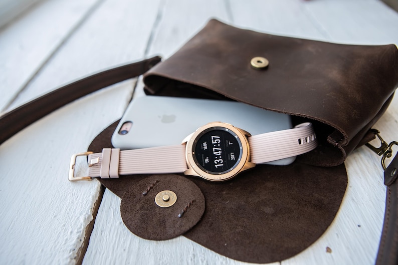 May include: A rose gold smartwatch with a light pink band rests on a white phone inside a brown leather crossbody bag. The watch displays the time 13:47:57. The bag is open, revealing its contents.