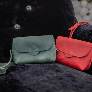 May include: Two leather crossbody bags in teal and red rest on a dark blue velvet surface. The teal bag has a matching strap, while the red bag has a red strap. Both bags feature a flap closure and a circular accent.