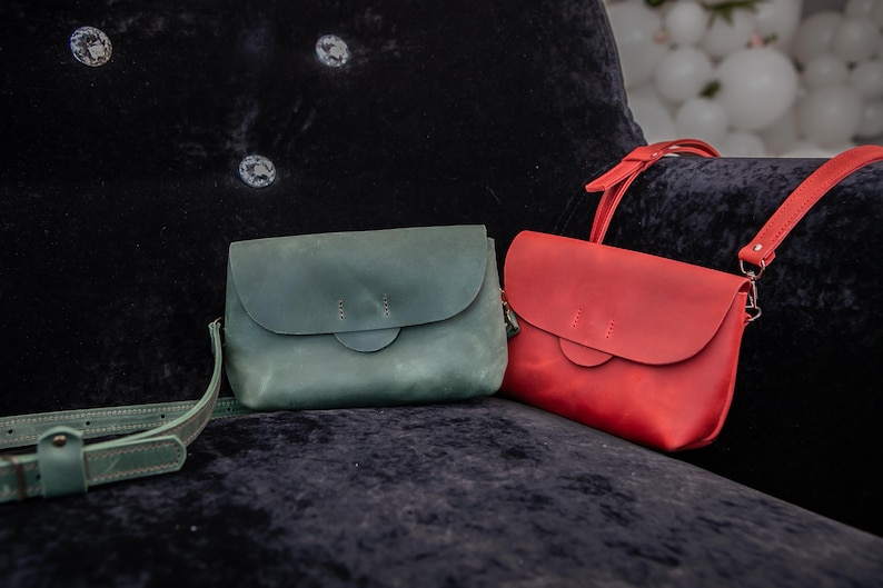 May include: Two leather crossbody bags in teal and red rest on a dark blue velvet surface. The teal bag has a matching strap, while the red bag has a red strap. Both bags feature a flap closure and a circular accent.