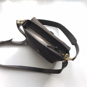 Puede incluir: Un bolso bandolera de cuero marr&oacute;n oscuro con una correa larga ajustable. El bolso tiene una cremallera y herrajes dorados. El bolso est&aacute; abierto, revelando el interior. El bolso est&aacute; dise&ntilde;ado para llevarse cruzado.