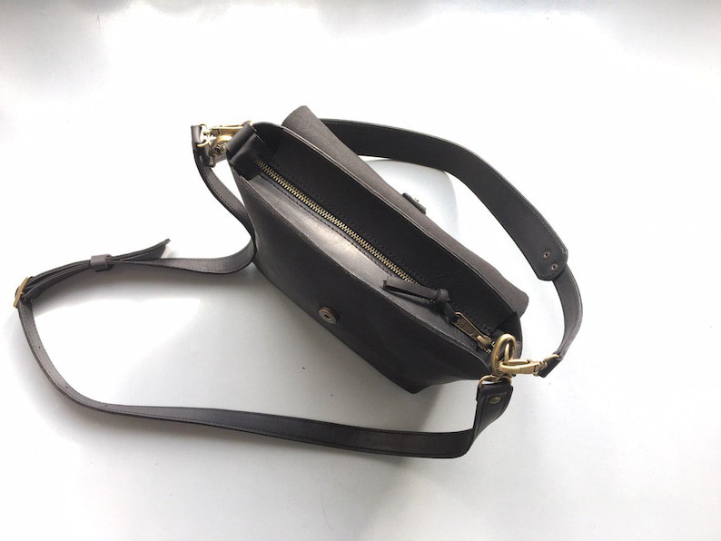 Puede incluir: Un bolso bandolera de cuero marr&oacute;n oscuro con una correa larga ajustable. El bolso tiene una cremallera y herrajes dorados. El bolso est&aacute; abierto, revelando el interior. El bolso est&aacute; dise&ntilde;ado para llevarse cruzado.