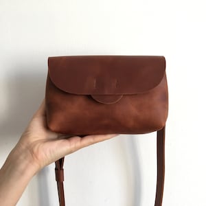May include: A brown leather crossbody bag with a rounded flap closure and a long, thin strap. The bag is held up by a hand against a white background. The bag has a simple, minimalist design.