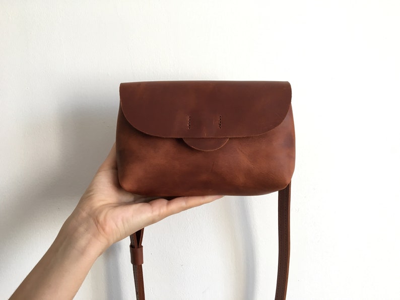 May include: A brown leather crossbody bag with a rounded flap closure and a long, thin strap. The bag is held up by a hand against a white background. The bag has a simple, minimalist design.
