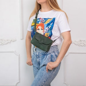 May include: A dark green leather crossbody bag with a flap closure and adjustable strap. The bag is worn over the shoulder. The person is wearing a white t-shirt with a colorful graphic print and blue jeans.