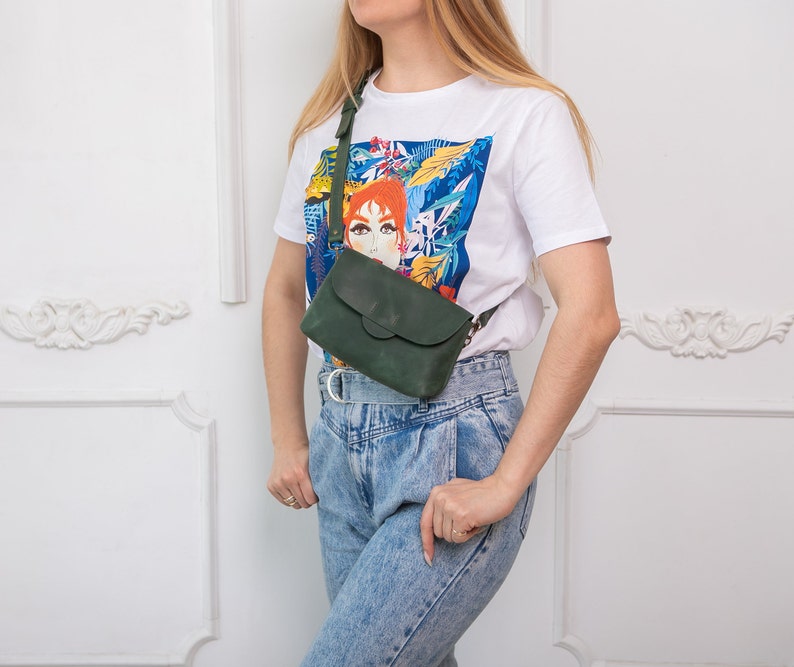 May include: A dark green leather crossbody bag with a flap closure and adjustable strap. The bag is worn over the shoulder. The person is wearing a white t-shirt with a colorful graphic print and blue jeans.
