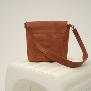 Puede incluir: Bolso bandolera de cuero marr&oacute;n con cierre de solapa y correa ajustable. El bolso tiene un bolsillo frontal y est&aacute; sobre un taburete blanco. El bolso tiene el texto "Blueberry Leatherworks" en la parte delantera.