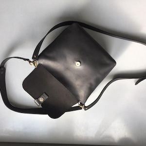 Puede incluir: Un bolso bandolera de cuero gris oscuro con cierre de solapa y correa ajustable. El bolso presenta un dise&ntilde;o cuadrado con un cierre y herrajes dorados. La correa es lo suficientemente larga para llevarla cruzada.