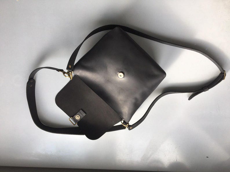 Puede incluir: Un bolso bandolera de cuero gris oscuro con cierre de solapa y correa ajustable. El bolso presenta un dise&ntilde;o cuadrado con un cierre y herrajes dorados. La correa es lo suficientemente larga para llevarla cruzada.