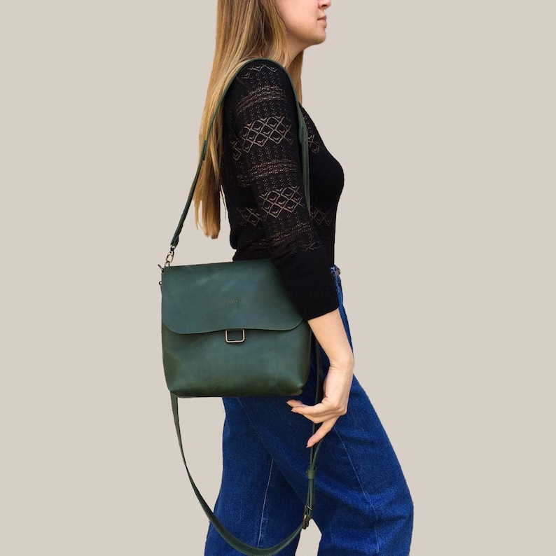 Puede incluir: Un bolso bandolera de cuero verde oscuro con cierre de solapa y correa ajustable para el hombro. El bolso tiene forma rectangular con una hebilla met&aacute;lica. El bolso se lleva al hombro.