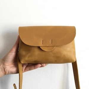 May include: A small, mustard-colored leather crossbody bag with a rounded flap closure. The bag has a simple design with a long, thin strap and is held by a hand against a white background. The bag's dimensions are approximately 8 inches wide and 6 inches tall.