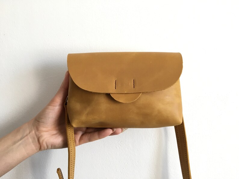 May include: A small, mustard-colored leather crossbody bag with a rounded flap closure. The bag has a simple design with a long, thin strap and is held by a hand against a white background. The bag's dimensions are approximately 8 inches wide and 6 inches tall.