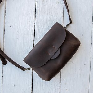 May include: A dark brown leather crossbody bag with a flap closure and adjustable strap. The bag is rectangular with a rounded flap and a circular clasp. The strap is attached with metal hardware. The bag is set against a white wooden background.