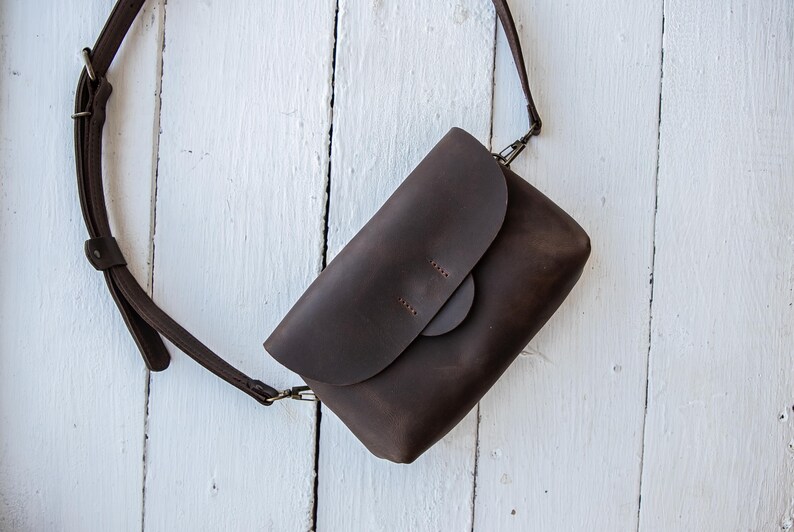 May include: A dark brown leather crossbody bag with a flap closure and adjustable strap. The bag is rectangular with a rounded flap and a circular clasp. The strap is attached with metal hardware. The bag is set against a white wooden background.