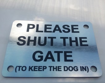 May include: A silver metal sign with black text that reads "Please Shut the Gate (To Keep the Dog In)". The sign has two holes in the top corners.