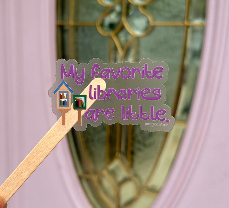 May include: A clear sticker with the text "My favourite libraries are little." and a small illustration of a little free library. The sticker is on a wooden stick.