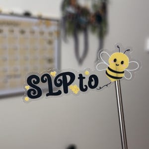May include: A clear sticker with the text "SLP to" and a cartoon bee with black and yellow stripes. The sticker is attached to a metal stick.