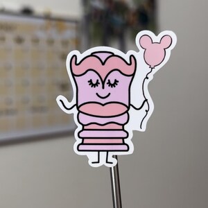 May include: A pink and white sticker of a cartoon character wearing a pink dress and holding a pink balloon with Mickey Mouse ears. The character has closed eyes and a smile.
