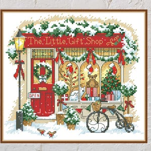 May include: Cross-stitch image of a festive gift shop. The shop front is red, adorned with wreaths, ribbons, and a Christmas tree. The sign above reads "The Little Gift Shop". Snow blankets the roof and the ground, creating a winter scene.