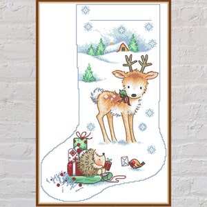 Peut inclure: Une chaussette de Noël blanche avec un cerf et un hérisson dans un paysage d'hiver enneigé. Le cerf est brun avec des bois et tient une branche de houx. Le hérisson est brun et porte un cadeau. Il y a un petit oiseau rouge en arrière-plan.