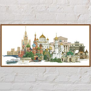 May include: A framed print depicting a detailed illustration of a cityscape, showcasing buildings such as churches with colourful domes and a tall tower. The artwork is set against a white background and framed in a brown wooden frame.