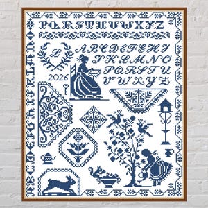 May include: A framed cross-stitch sampler in navy blue, featuring an alphabet, decorative borders, and various motifs like a woman, birds, and a rabbit. The design includes the year 2026 and a variety of floral and geometric patterns.