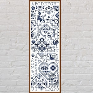 May include: A framed cross-stitch sampler in navy blue, featuring floral and geometric patterns, letters, and numbers. The design includes a heart, birds, and other decorative elements. The sampler is displayed against a white brick wall.