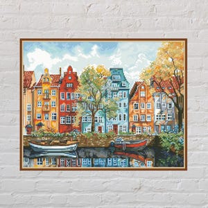 May include: A framed watercolour painting depicting a canal scene with colourful buildings. The buildings have red, orange, blue, and green facades. Boats are in the water, with reflections. Trees with autumnal leaves frame the scene.