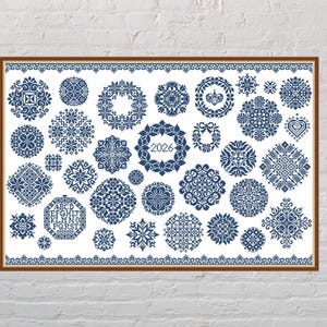 Puede incluir: Obra de arte enmarcada con una colección de diseños de punto de cruz azules y blancos. Los diseños incluyen patrones florales, formas geométricas y el año "2026". La obra está enmarcada en un marco de madera marrón y se exhibe contra una pared de ladrillos blancos.