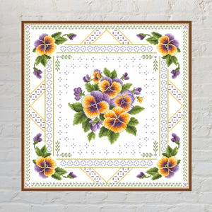 May include: A square cross-stitch pattern featuring a central bouquet of purple and yellow pansies with green leaves. The design is framed by a geometric border of purple and yellow stitches on a white background, enclosed in a brown frame.
