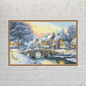 May include: A rectangular wall art print featuring a winter landscape with a stone bridge over a stream, snow-covered trees, and a cluster of houses with glowing windows under a soft yellow and blue sky, all framed by a thin brown border.