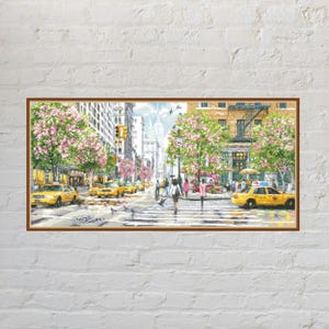 Op de afbeelding: Een rechthoekig ingelijst schilderij met een straatbeeld van New York City met gele taxi's, voetgangers op een zebrapad en bomen met roze bloesems. Hoge gebouwen staan langs de straat onder een heldere lucht met een duidelijk zicht op de stad.