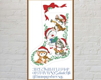 May include: A cross-stitch pattern of a Christmas stocking featuring four kittens wearing red Santa hats, with one playing a trumpet, one singing from a book, one holding cookies, and one playing a violin. The alphabet is printed below in blue.