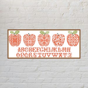 May include: A rectangular cross-stitch sampler featuring five stylised orange apples with intricate geometric patterns and green leaves, positioned above two rows of the English alphabet in a decorative orange font, all enclosed in a brown frame.