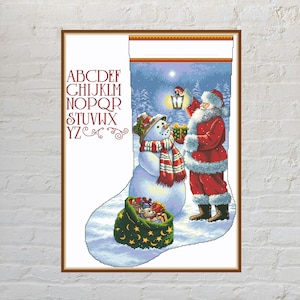 Può includere: Uno schema a punto croce per calza natalizia con l'alfabeto in lettere rosse sulla sinistra. L'immagine principale mostra Babbo Natale con una lanterna e un regalo accanto a un pupazzo di neve con sciarpa e un sacco verde di giocattoli.