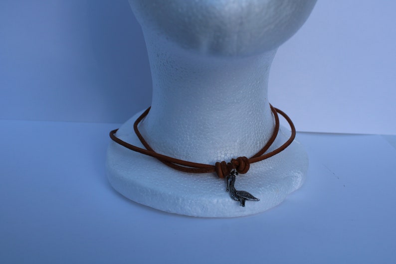 Może przedstawiać: Brązowy sk&oacute;rzany naszyjnik ze srebrnym wisiorkiem w kształcie ptaka.