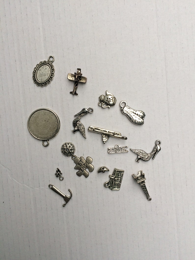 Może przedstawiać: Kolekcja srebrnych metalowych zawieszek, w tym samolot, czajnik, ptak, element układanki, piłka golfowa, młotek, książka, wieża Eiffla i kilka innych abstrakcyjnych kształt&oacute;w.